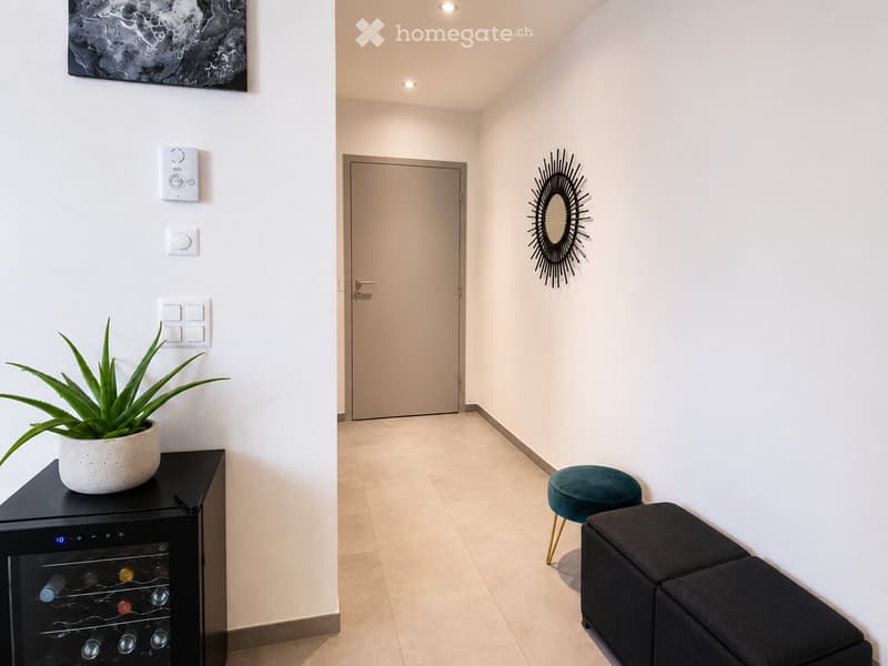 Modern hallway with white walls, beige tiled floor, a closed door, a small black bench, a teal stool, a wall-mounted mirror with a decorative frame, a potted plant on a black side table, and a wine cooler with multiple bottles.