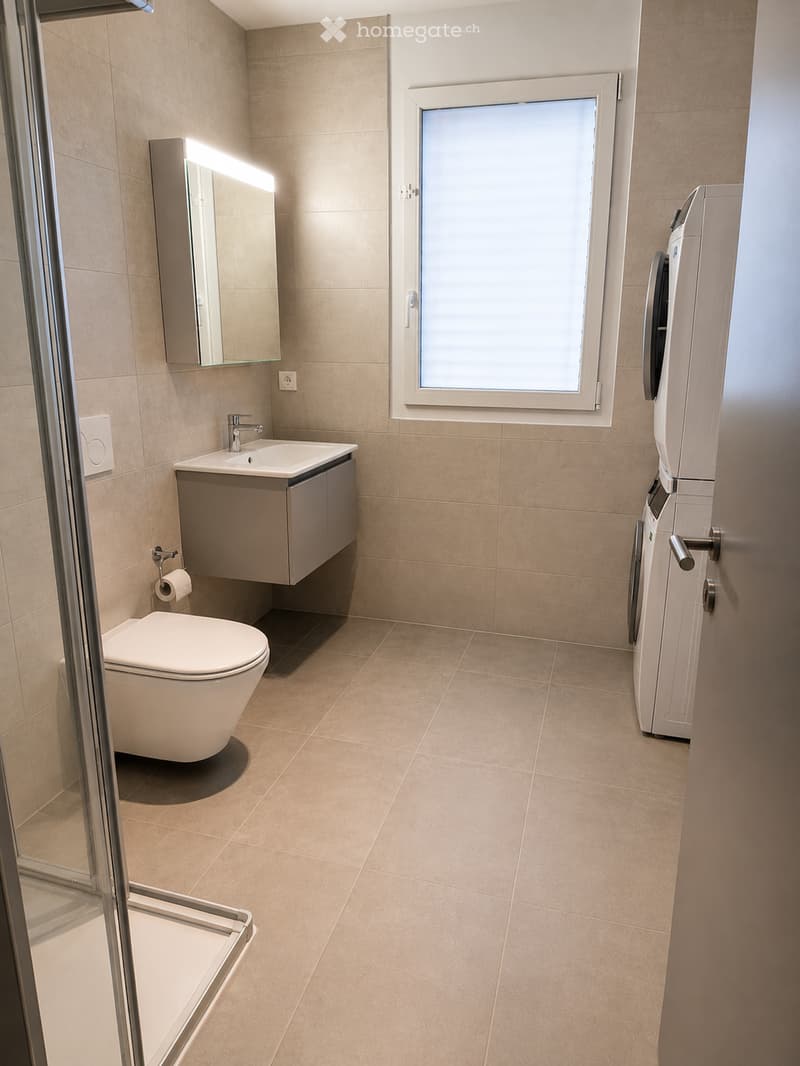 Modern bathroom with beige tiles, a toilet, a sink with a cabinet, a mirror, and a washing machine in the corner.