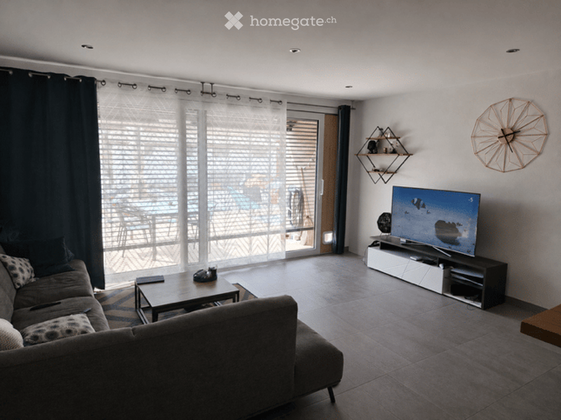 Modern living room with large sliding glass doors leading to a balcony, grey sectional sofa, coffee table, flat-screen TV on a stand, wall-mounted shelves, and a decorative wall clock.