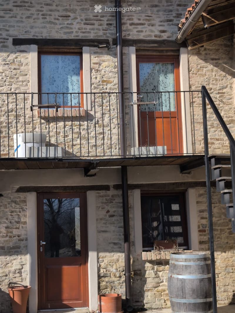 Stone building with a balcony, wooden doors, and a metal railing. Features include a large barrel and potted plants near the entrance.