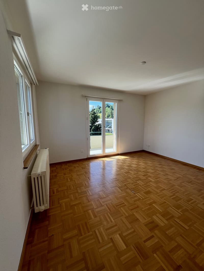 Empty room with wooden parquet flooring, white walls, large windows, and a sliding glass door leading to a balcony.