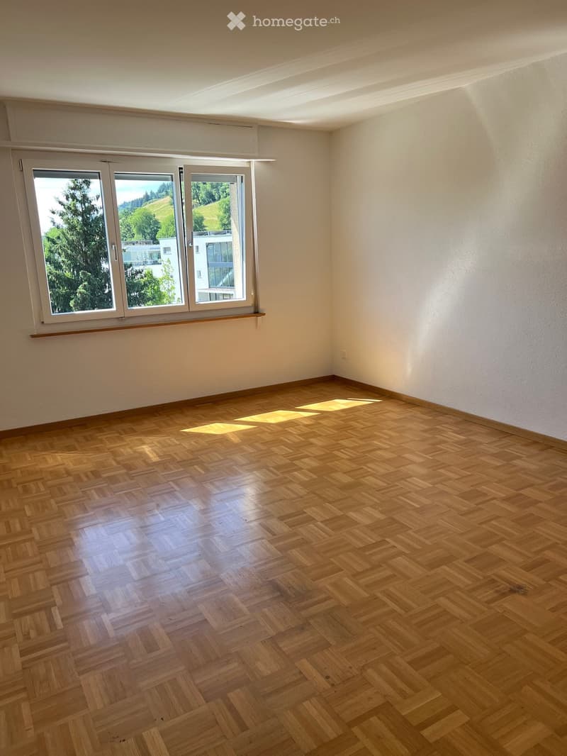Empty room with wooden herringbone flooring and large windows offering a view of greenery and buildings outside.
