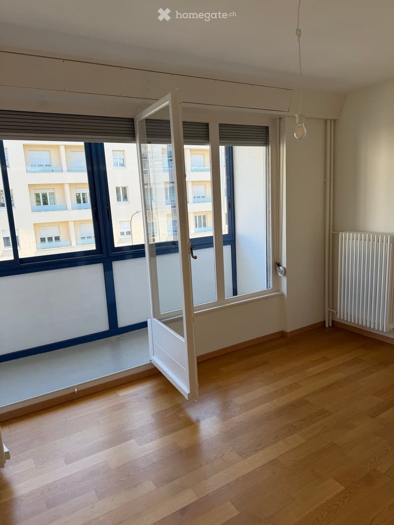 Empty room with wooden flooring, large windows with open shutters, radiator, and visible light fixture.