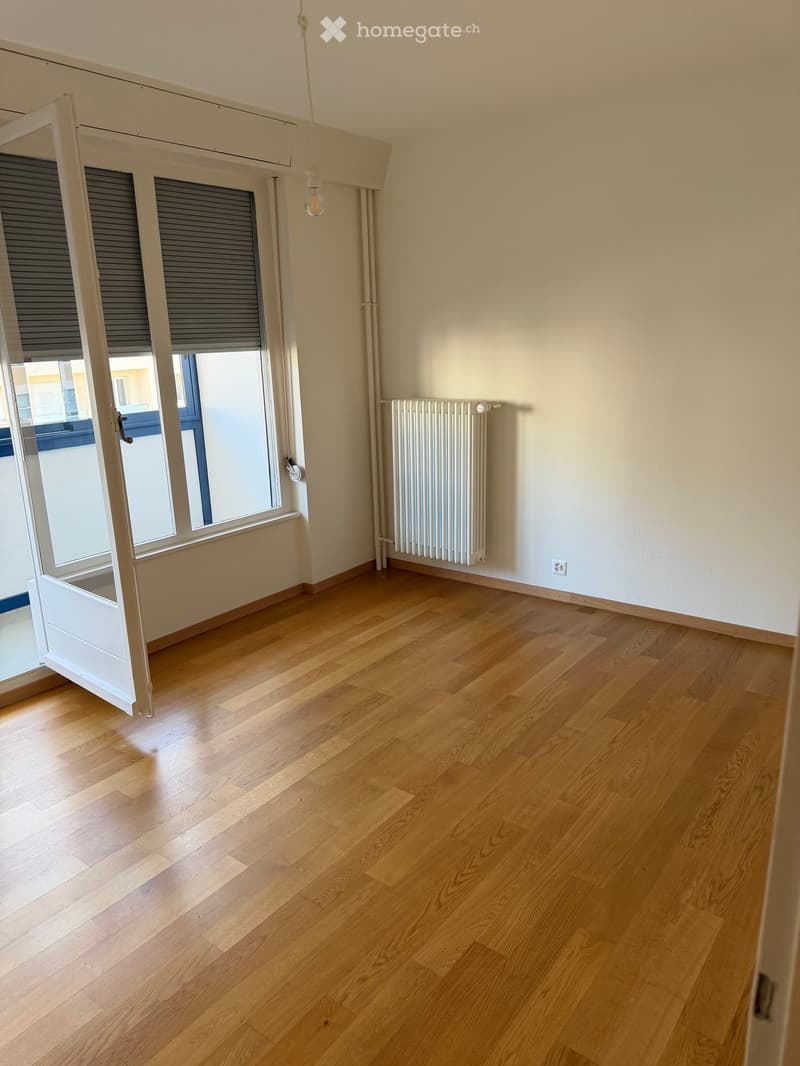 Empty room with wooden flooring, white walls, radiator, and large window with blinds.