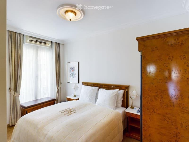 Elegant bedroom with a large wooden bed, white bedding, two nightstands with lamps, a bench at the foot of the bed, and a large wooden wardrobe.