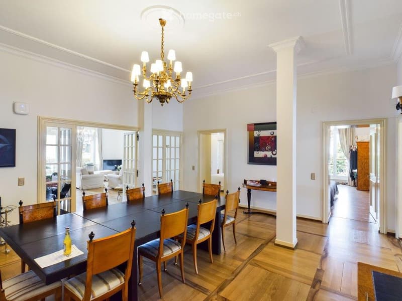 Elegant dining room with a large wooden dining table surrounded by upholstered chairs, a chandelier hanging from the ceiling, and a painting on the wall. The room features wooden flooring and opens to a living area with a sofa and television.