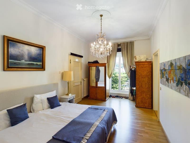 Elegant bedroom with a large bed, nightstand, lamp, wardrobe, chandelier, and artwork on the walls. The room features wooden flooring and large windows with curtains.