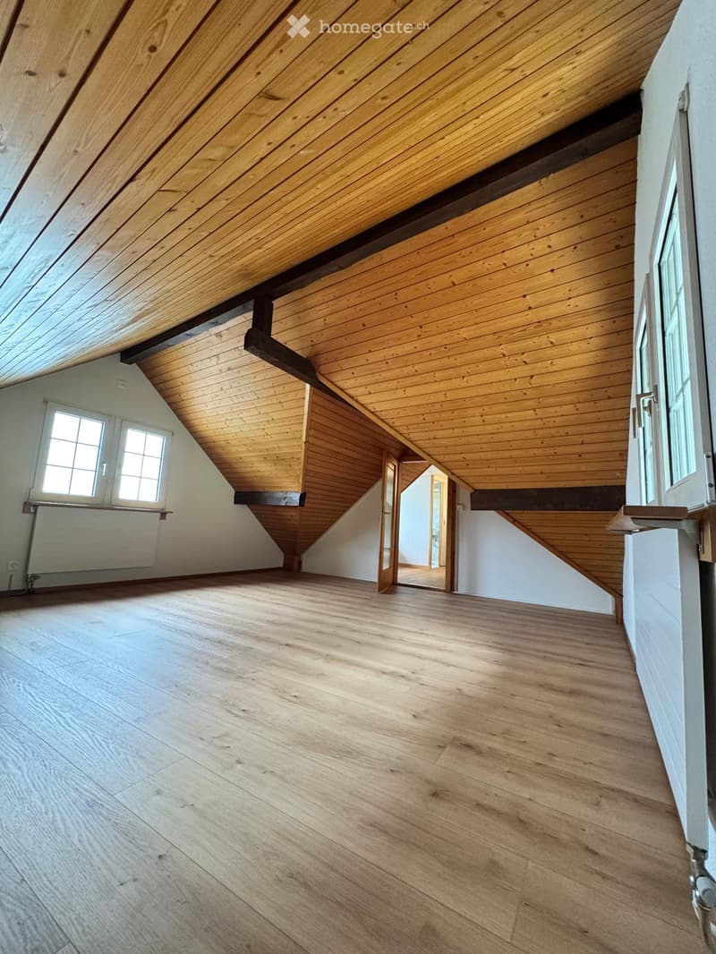 Empty attic space with wooden floors and slanted wooden ceilings, featuring large windows and open doorways.