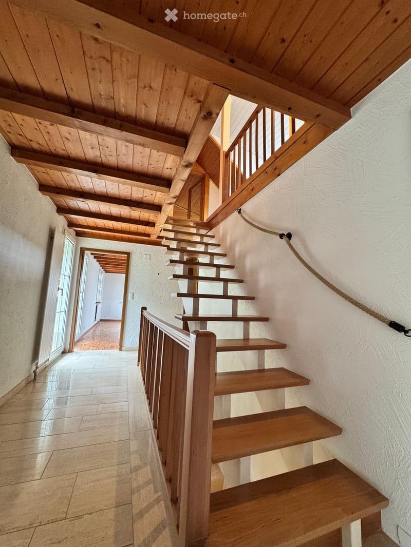 Wooden staircase with a wooden railing and exposed wooden beams on the ceiling, leading to an open area with tiled flooring and white walls.