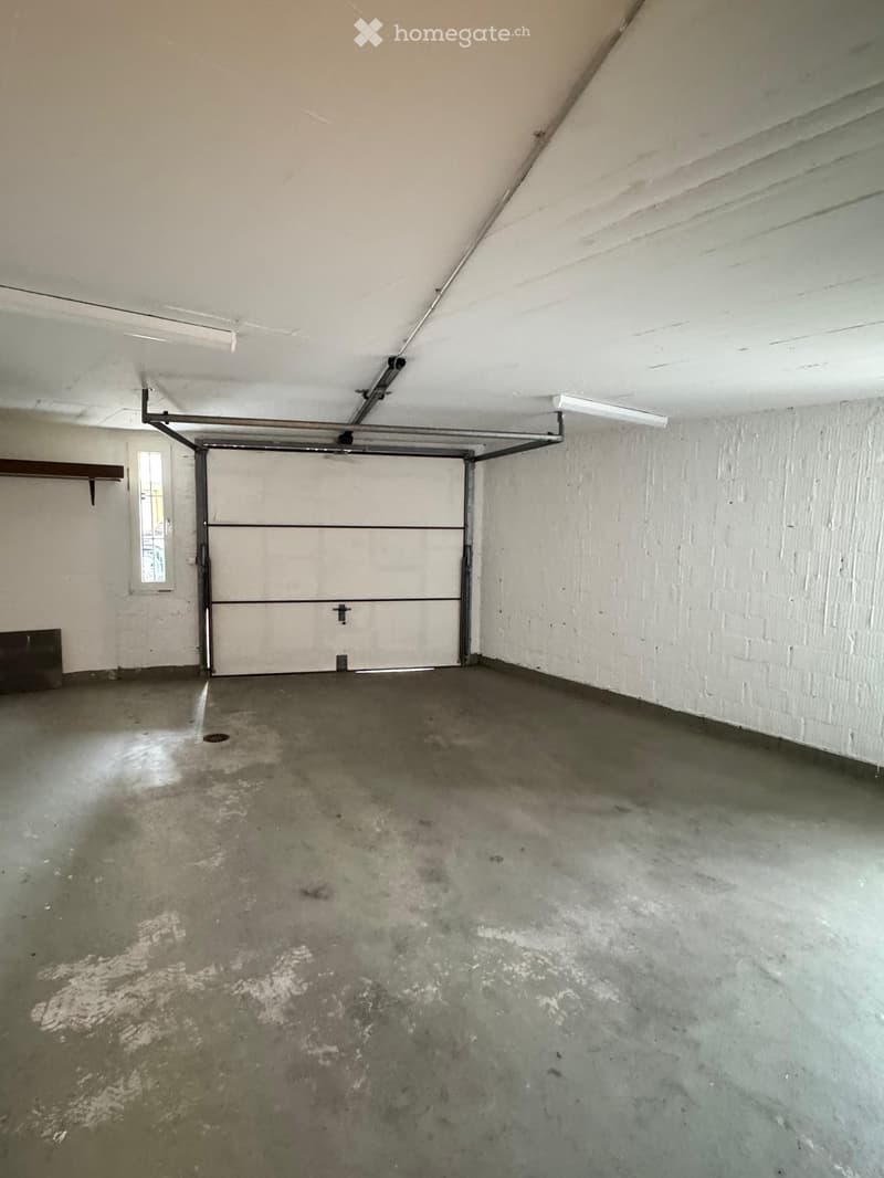 Empty garage with a concrete floor, white brick walls, and a large overhead door. There is a small window on the left side and a single light fixture on the ceiling.