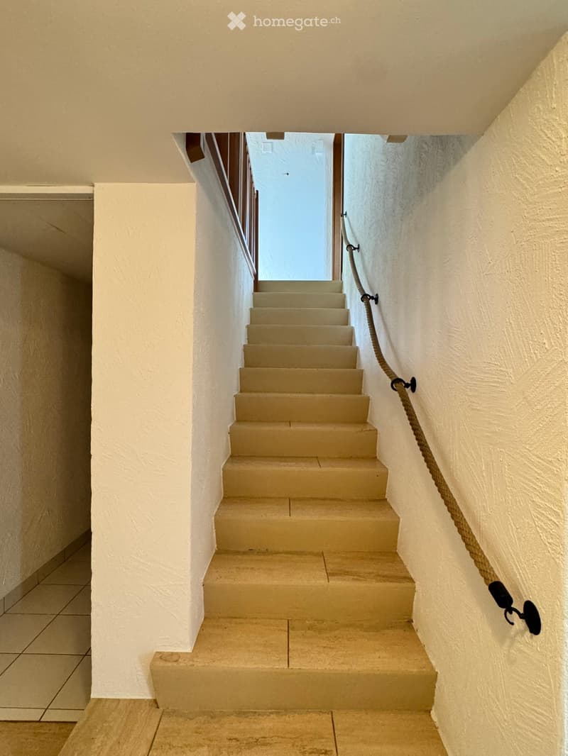A narrow staircase with beige steps and white walls, featuring a rope handrail on one side and a wooden handrail on the other, leading to a bright opening at the top.