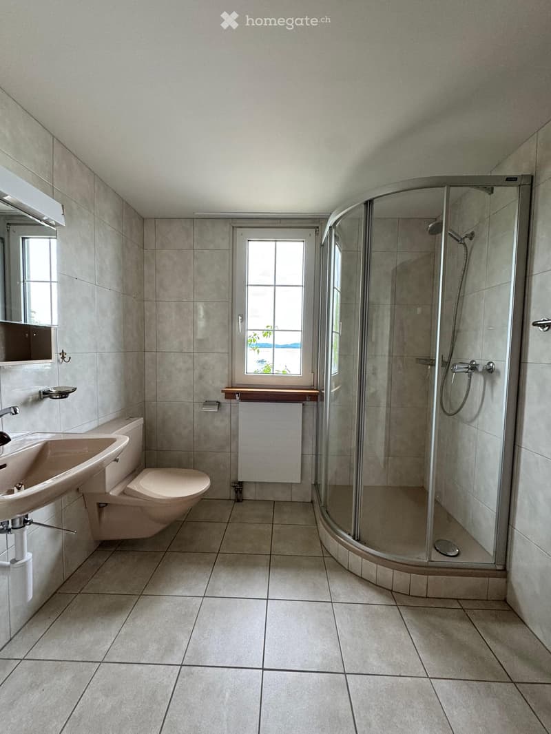 Modern bathroom with tiled walls and floor, featuring a sink, toilet, glass-enclosed shower, and a window with a view.