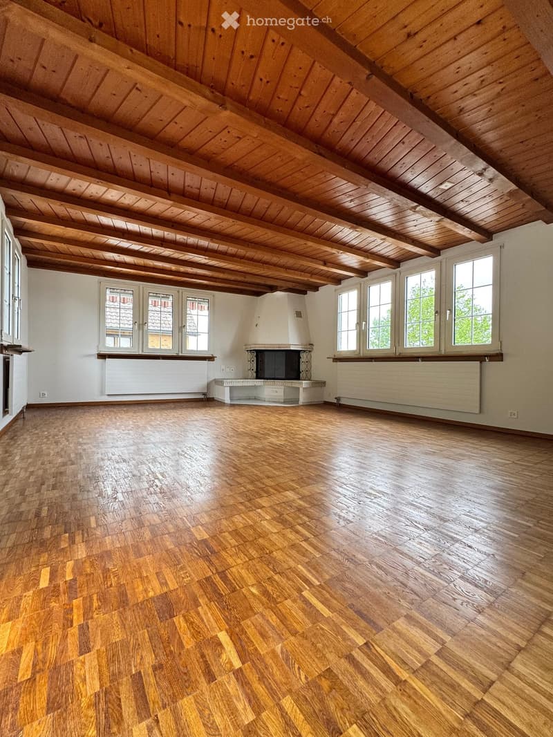 Empty room with wooden floors, exposed wooden ceiling beams, large windows, and a fireplace.