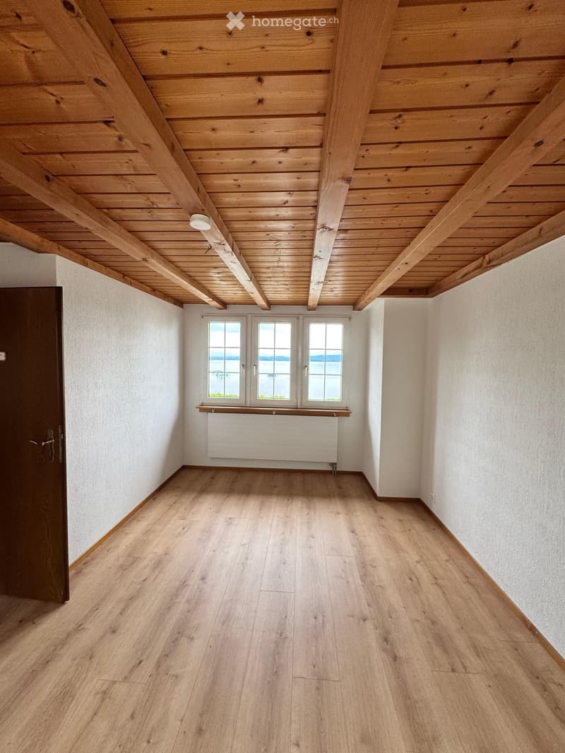 Empty room with wooden flooring, white walls, exposed wooden ceiling beams, and a window with a view of the water.