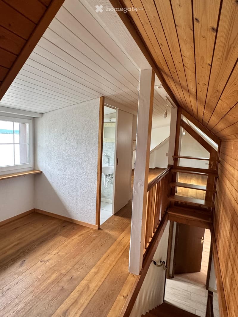 Wooden staircase leading to an open area with a small bathroom visible at the end. The hallway has wooden flooring and white walls, with a window on the left side providing natural light.
