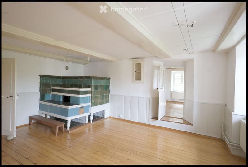 Bright room with wooden flooring, white walls, and a fireplace with green and blue tiles. A small bench is placed in front of the fireplace.