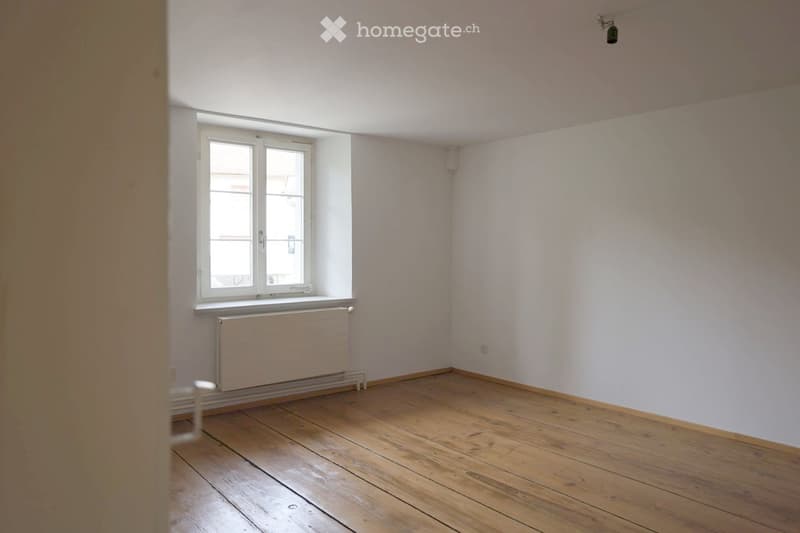 Empty room with wooden flooring, white walls, a window with white frame, and a radiator below the window.