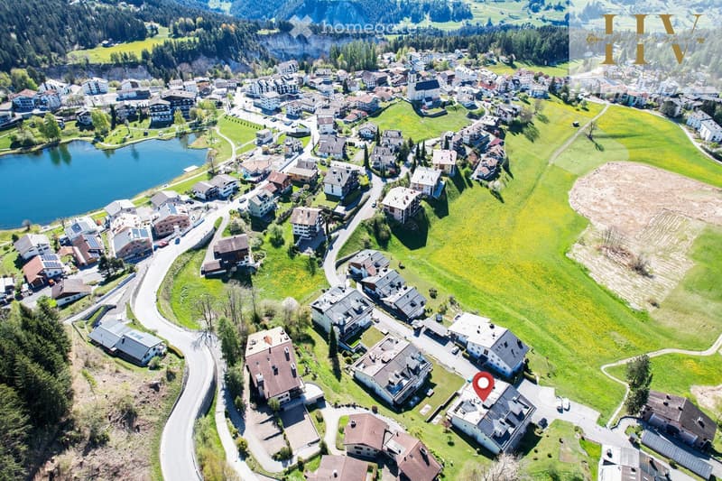 Aerial view of a picturesque village with numerous houses, green fields, a lake, and surrounding mountains.
