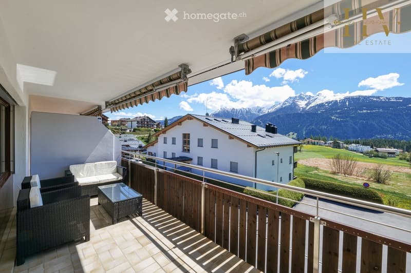 Balcony with a view of mountains and a village, featuring a sofa, a table, and a chair.