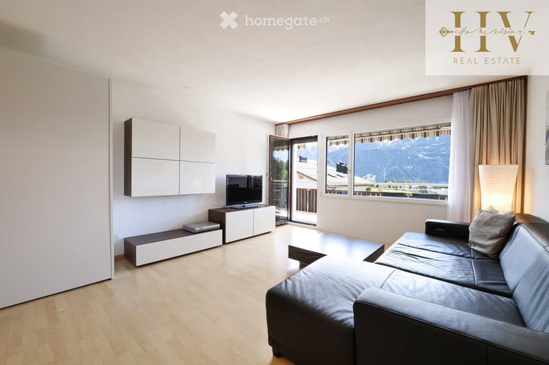 Modern living room with large windows offering a mountain view, featuring a black leather sofa, a coffee table, a TV stand, and white cabinets.