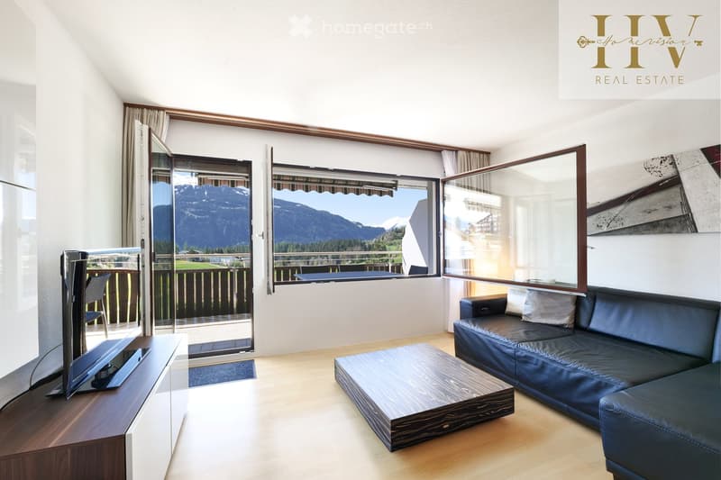 Modern living room with large windows offering a scenic mountain view, featuring a dark leather sofa, wooden coffee table, flat-screen TV on a wooden stand, and a chair near the balcony.