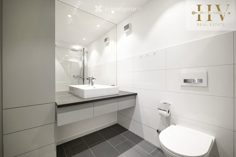 Modern bathroom with white tiles, a large sink, a toilet, a shower area, and a mirror. The space features sleek, contemporary design elements.