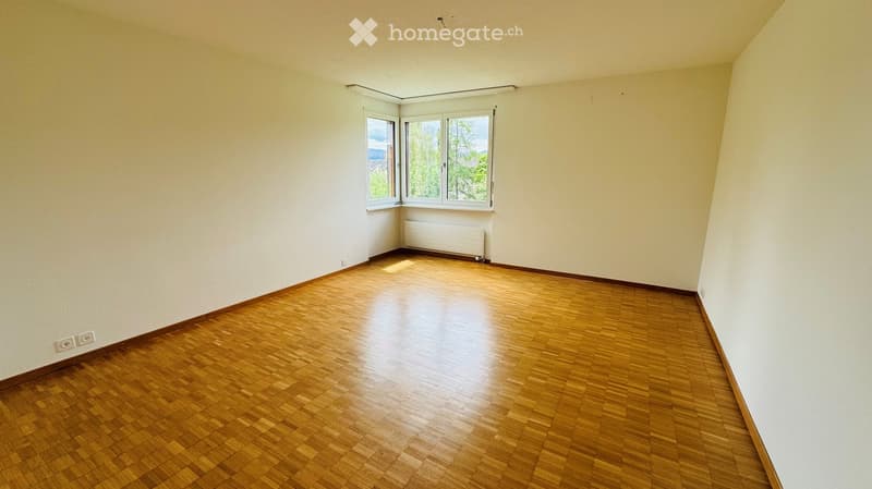 Empty room with wooden flooring, white walls, and large windows offering a view of trees and a distant landscape.