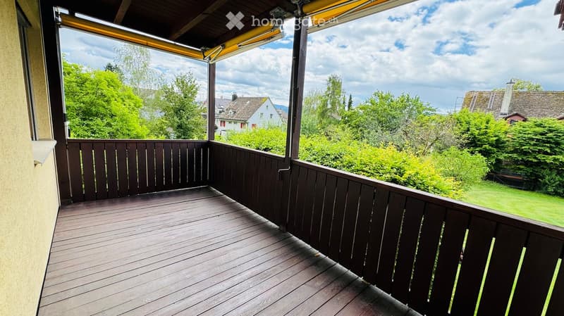 Wooden balcony with dark railing, retractable awning, overlooking a green garden and houses.