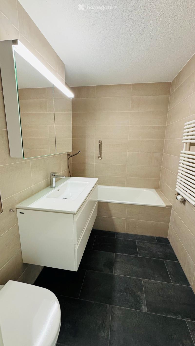 Modern bathroom with a white sink, bathtub, radiator, and mirror. The walls are tiled in beige, and the floor is dark grey.