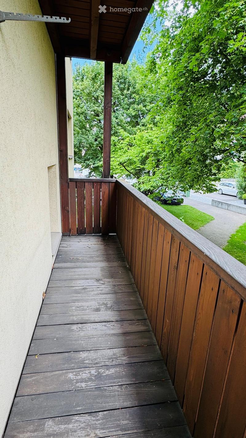 Wooden balcony with railing, attached to a building, overlooking a green area with trees and parked cars.