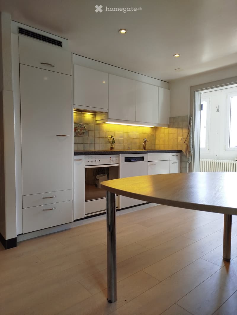 Modern kitchen with white cabinets, wooden floor, central table, built-in oven and dishwasher, and a window with natural light.