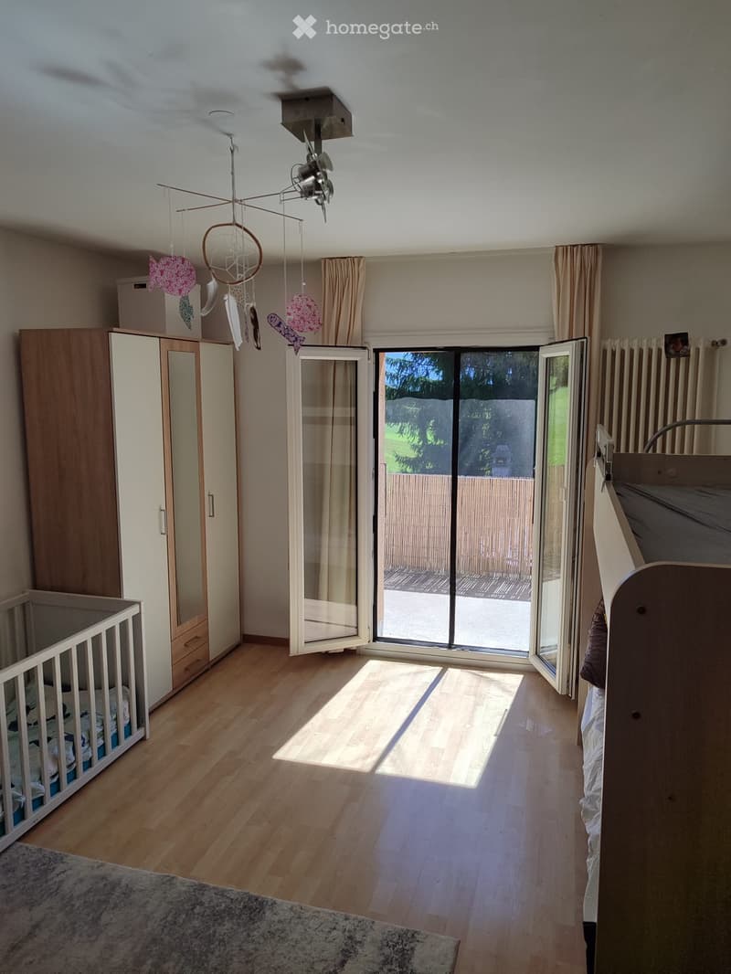 Brightly lit bedroom with wooden flooring, large sliding glass doors leading to a patio, a wardrobe, a crib, a bed, and a ceiling fan with hanging toys.