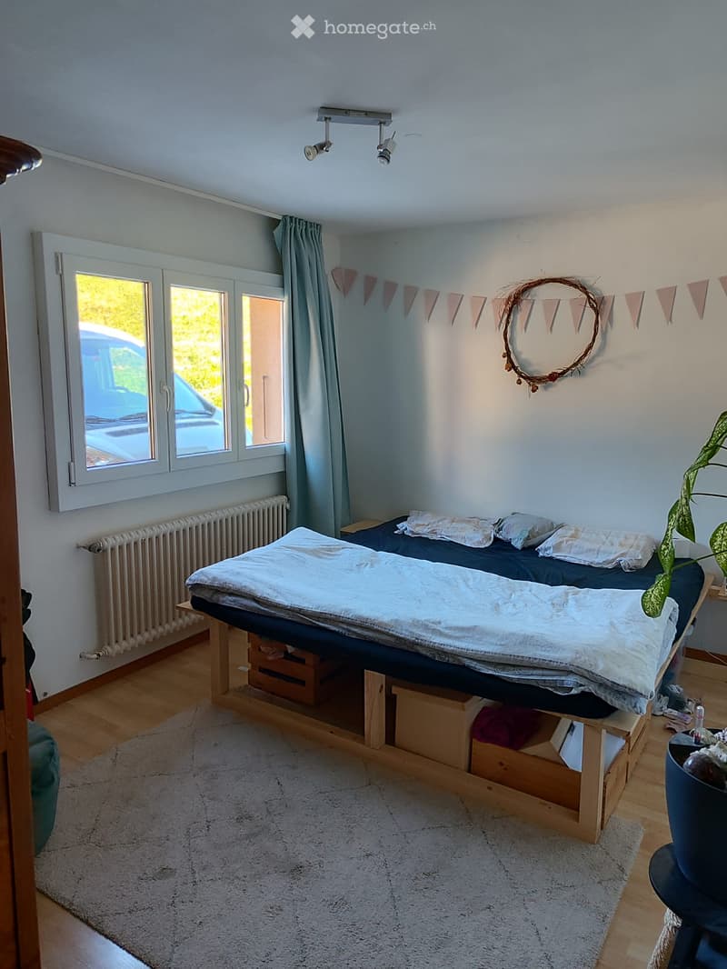 Cozy bedroom with a raised wooden bed, pillows, blanket, nightstand, plant, and decorative wreath on the wall. The room features large windows with curtains, a radiator, and a soft white rug on the floor.