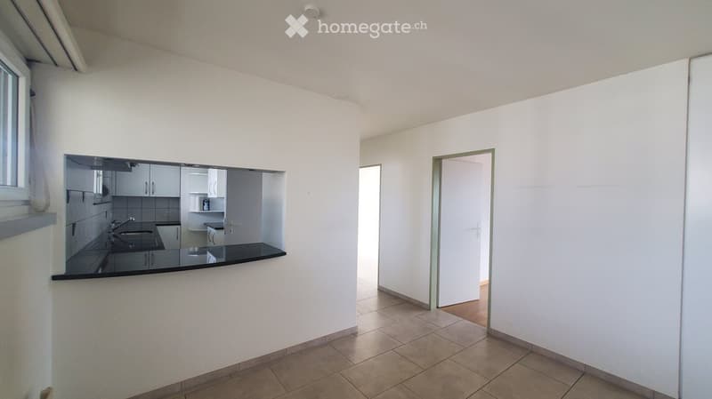 Empty room with white walls, tiled floor, and a doorway leading to another space. A mirror on one wall reflects a small kitchen area with cabinets, sink, and countertop.