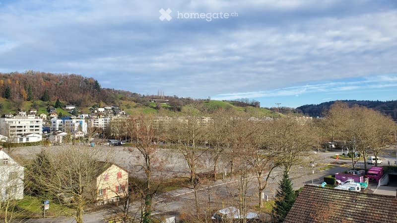 A panoramic view of a suburban area with residential buildings, trees, and a parking lot, surrounded by hills and a partly cloudy sky.