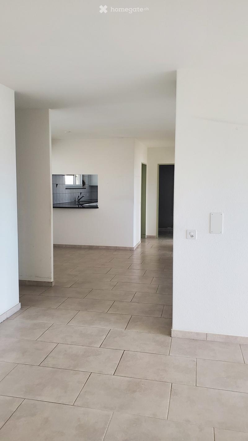 Empty hallway with tiled floor and white walls, leading to a small kitchen area with sink and cabinets.