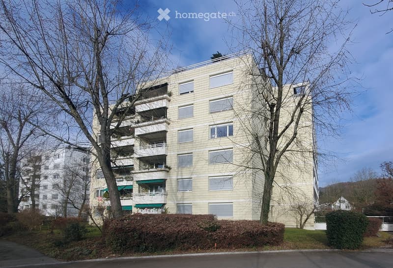 Modern multi-story residential building with light-colored facade, multiple balconies, and large windows. Surrounded by trees and greenery.