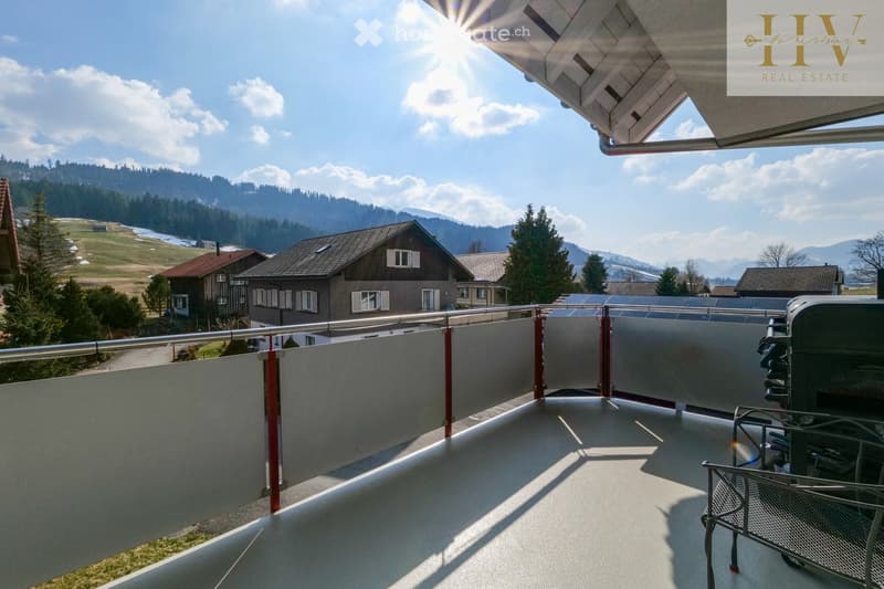 Balcony with a modern railing, overlooking a scenic countryside with houses and mountains in the background.
