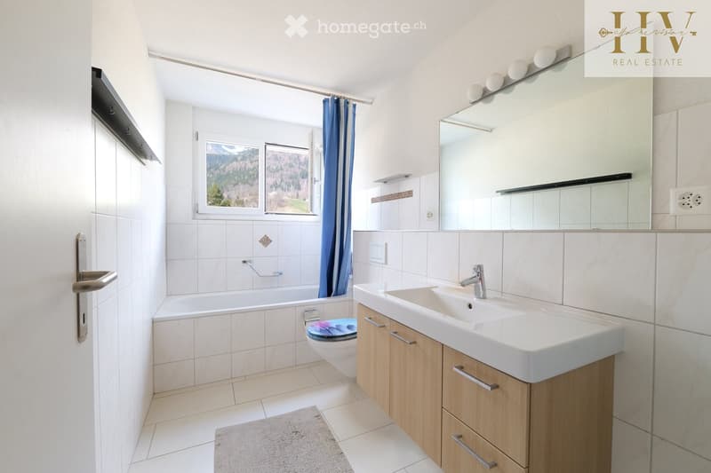 Modern bathroom with white tiles, a bathtub, a toilet, a sink with wooden cabinetry, and a large mirror with integrated lighting. The room features a window with blue curtains offering a scenic mountain view.