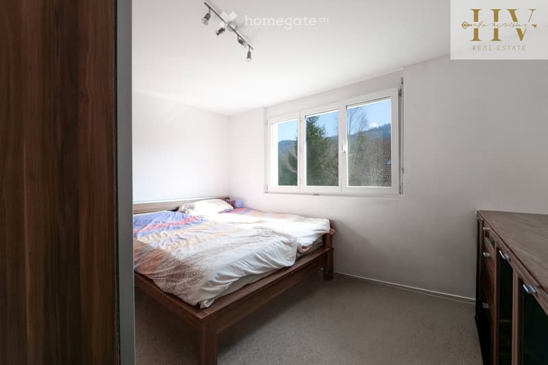 Bright bedroom with a wooden bed, colorful bedding, a nightstand, and a wardrobe. Large windows offer a scenic view.