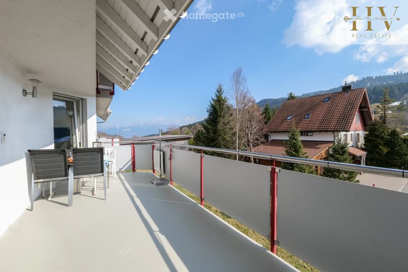 Balcony with white floor, metal railing, and outdoor furniture including chairs and a table, overlooking a scenic mountain landscape.