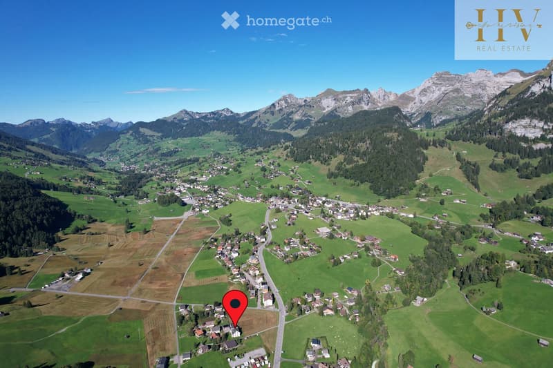 Aerial view of a picturesque village nestled in a lush green valley surrounded by mountains. The village features scattered houses, roads, and fields, with a red marker indicating a specific location.