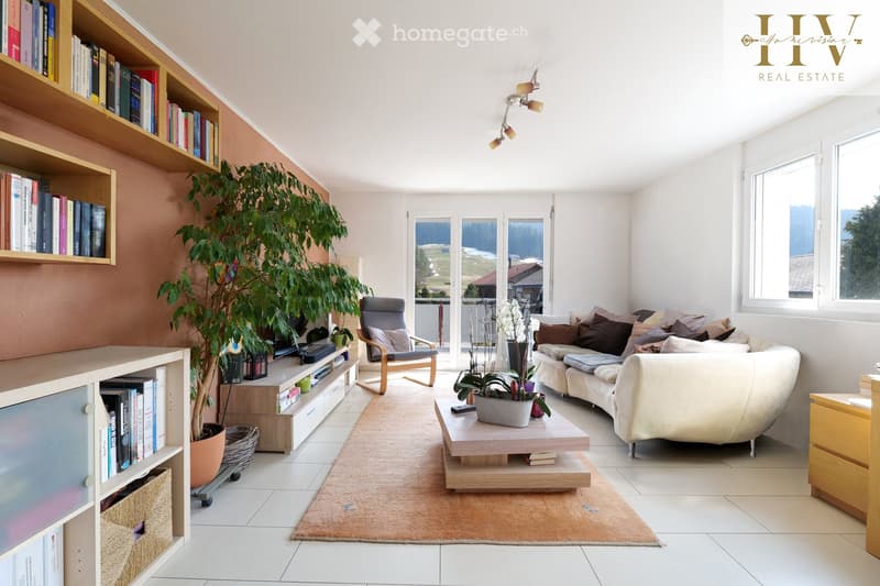 Bright living room with large windows offering a scenic view, featuring a white sofa, wooden coffee table, armchair, bookshelf, and potted plants.