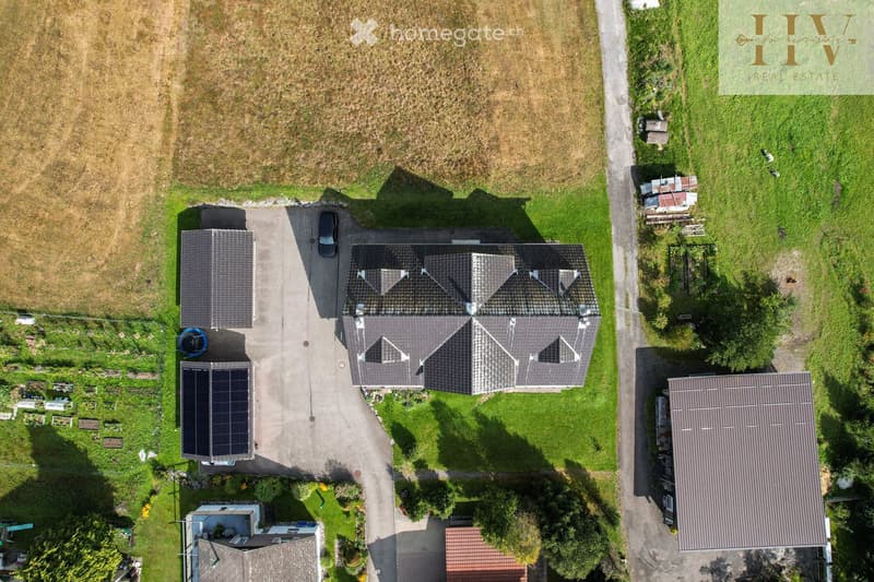 Aerial view of a two-story house with a grey roof, surrounded by a well-maintained lawn, driveway, and neighboring structures.