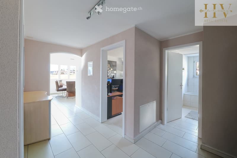A well-lit hallway with white tiled flooring, beige walls, and several doors leading to different rooms. The hallway features a wooden reception desk on the left and a small seating area with chairs and a table visible through an open doorway on the left