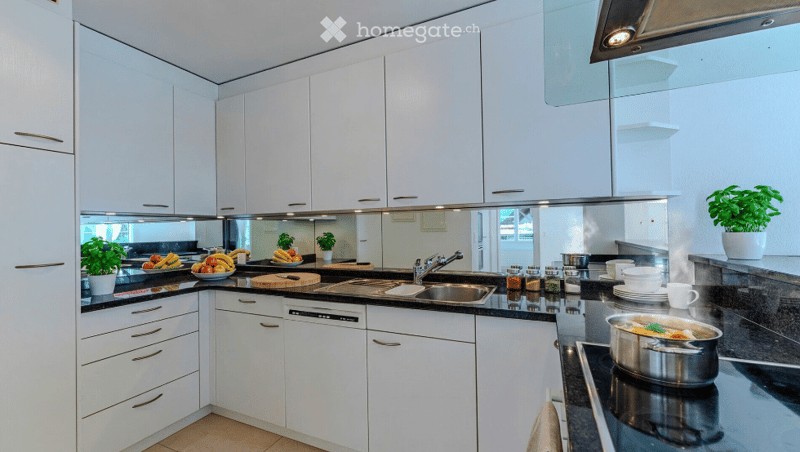 Modern kitchen with white cabinets, stainless steel appliances, and a black countertop. Features a sink, stove, and various kitchen items including a pot, cutting board, and potted plants.