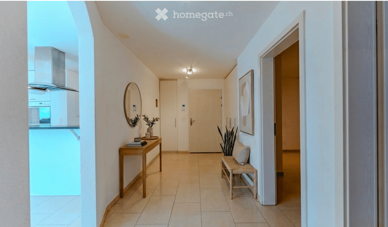 Bright hallway with tiled floor, white walls, and recessed lighting. Features a small wooden bench, a small table with books and a plant, and a round mirror on the wall.