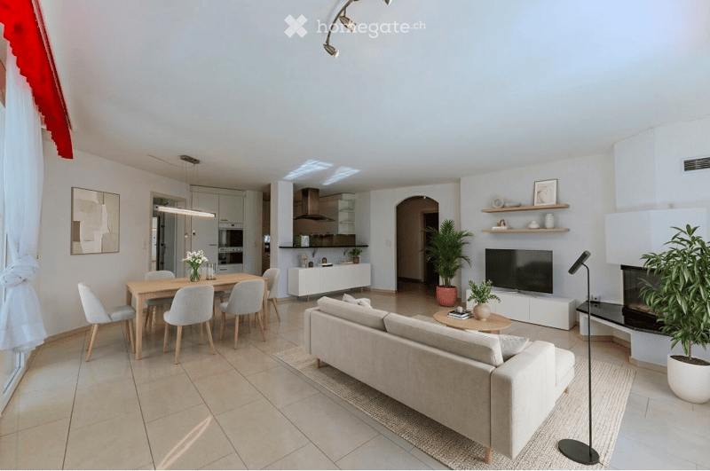 Modern open-plan living and dining area with a light-colored sofa, coffee table, dining table with chairs, and a fireplace. The space features a kitchen area with white cabinets and stainless steel appliances, along with decorative shelves and potted plan