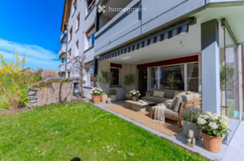 Well-maintained garden area with a wooden deck, outdoor seating, potted plants, and a view of a modern building.