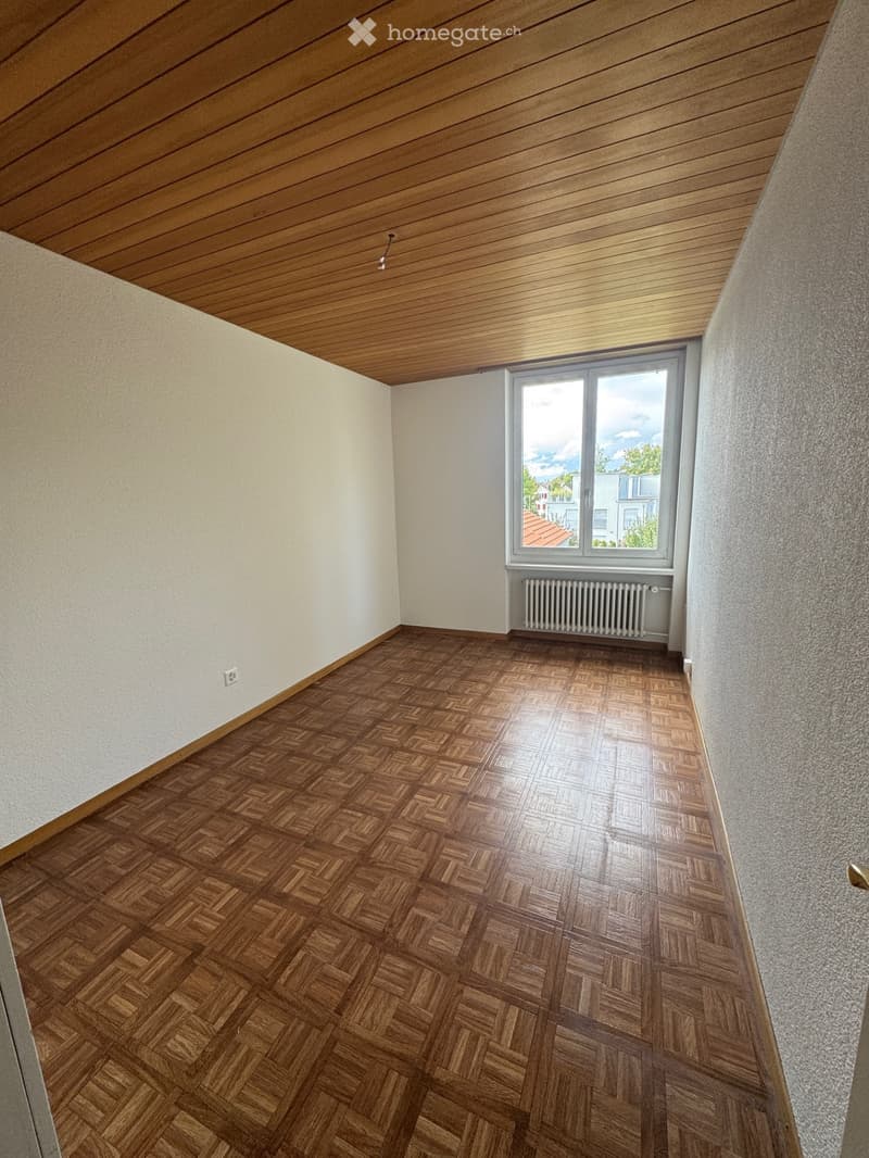 Empty room with wooden herringbone flooring, white walls, and a large window with a radiator below it. The ceiling is wooden with a slanted design.